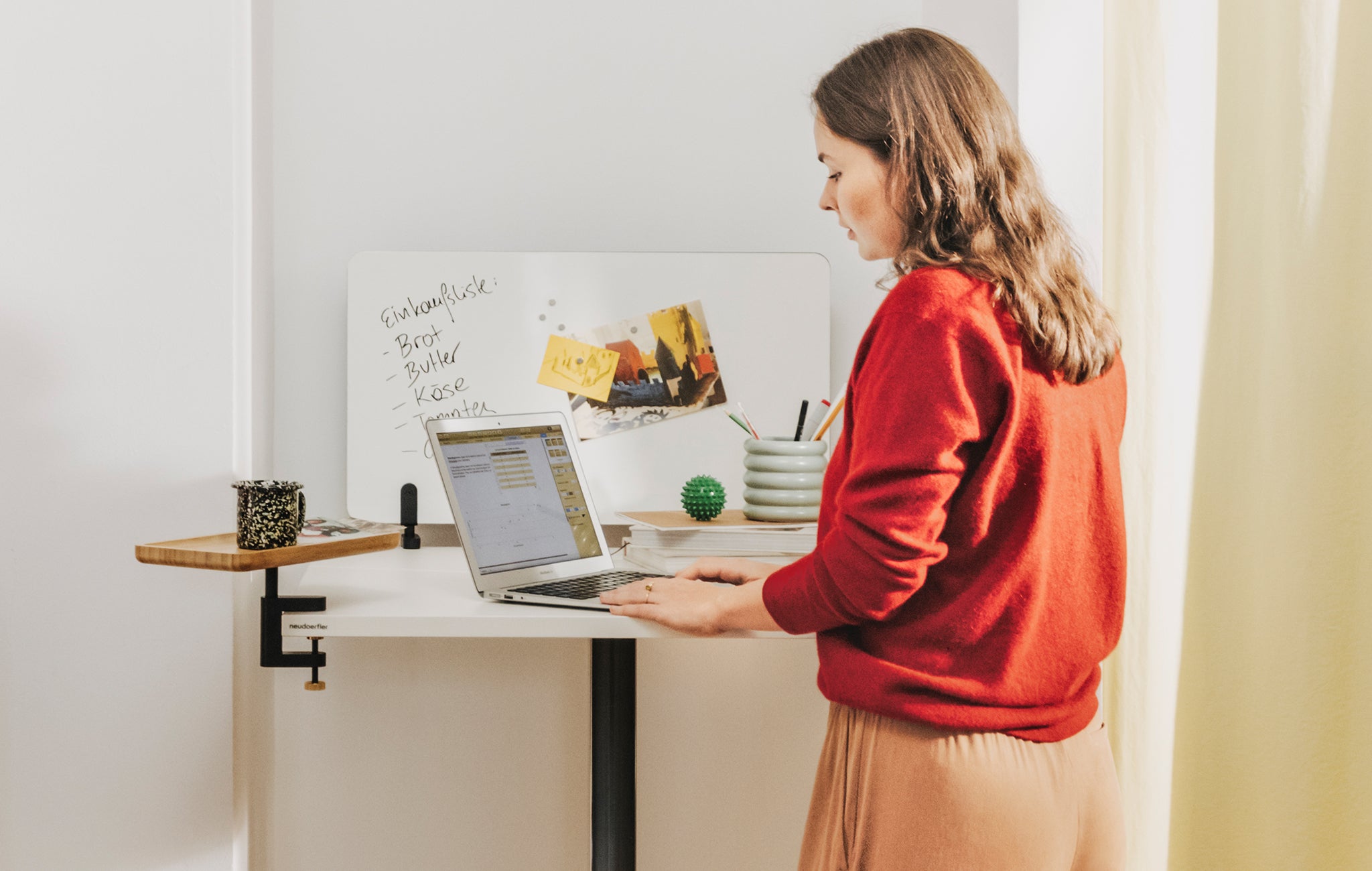  Eine junge Frau mit braunen, langen Haaren und einem roten Pullover arbeitet im Stehen auf einem Laptop, der auf einem weißen Move Mini Arbeitstisch steht. Auf dem Tisch ist ein Whiteboard, das beschriftet ist, montiert und ein Anbautablet aus Holz, auf dem eine Tasse steht. 