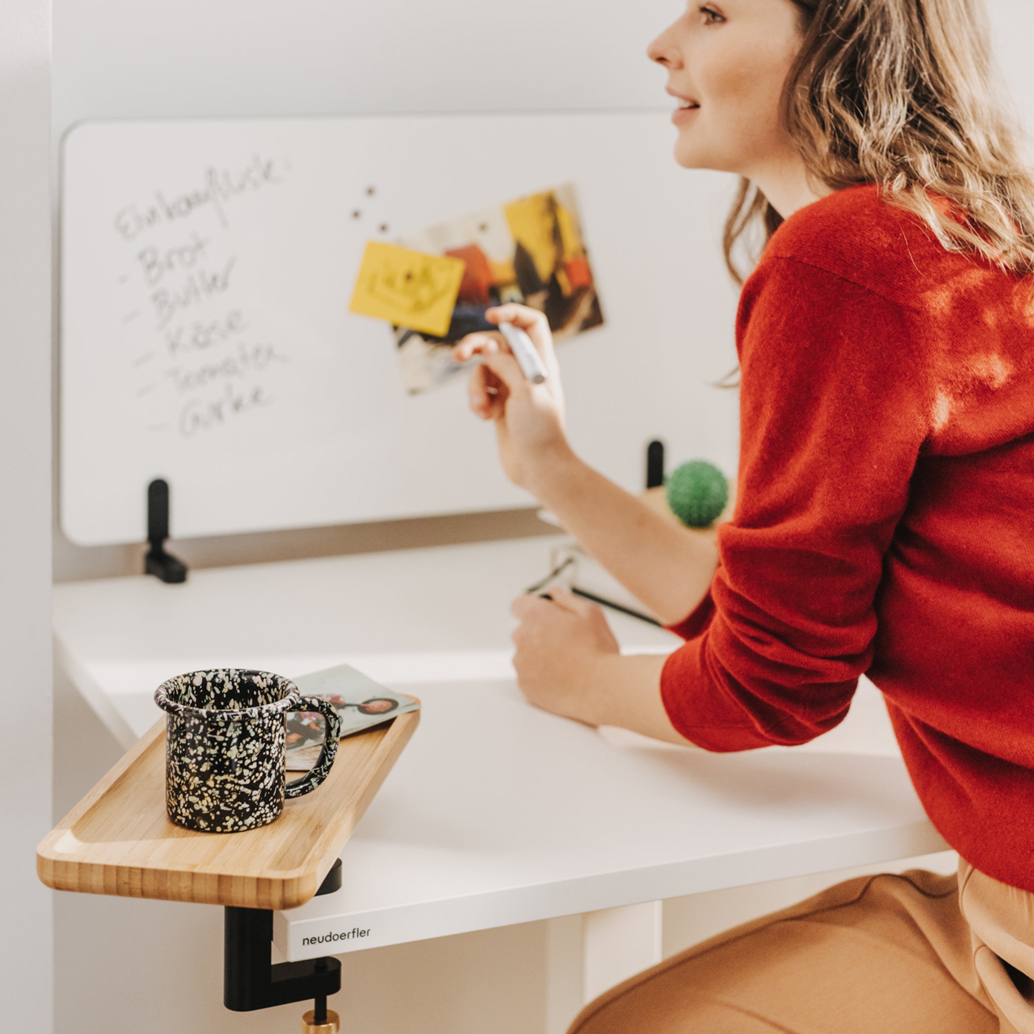 Eine junge Frau mit braunen, langen Haaren und einem roten Pullover sitzt bei einem weißen Move Mini Arbeitstisch. Auf dem Tisch ist ein Whiteboard, das beschriftet ist, montiert und ein Anbautablet aus Holz, auf dem eine Tasse steht. Sie hält einen Stift in der Hand.