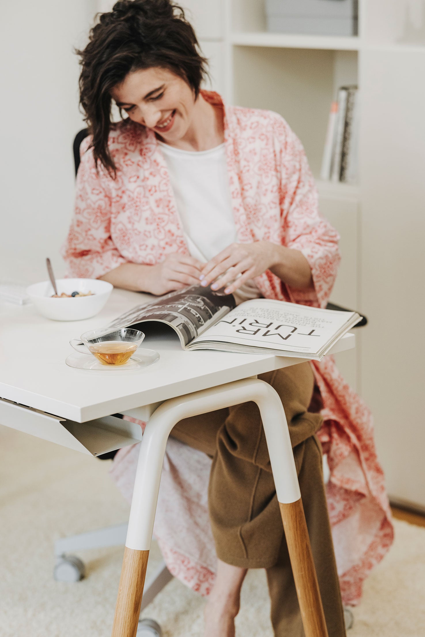 Man sieht einen Arbeitsplatz mit einem weißen Arbeitstisch, der Tischbeine aus Holz hat. Eine Frau sitzt davor und lächelt, während sie ein Magazin durchblättert.