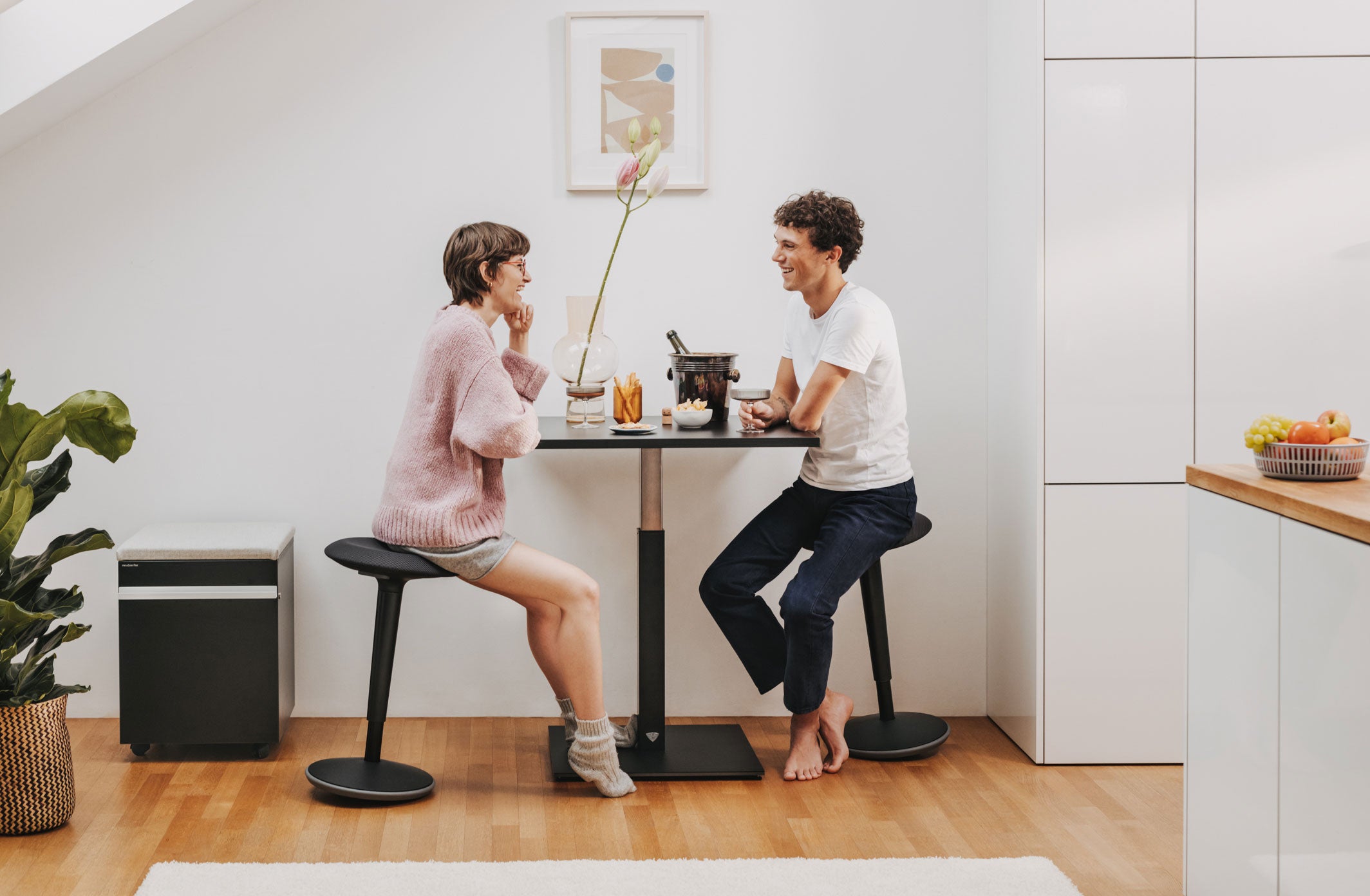 Eine Frau und ein Mann sitzen mit Hockern an einem Stehtisch mit Snacks und Drinks. Rechts ist eine Küche zu sehen und links ein schwarzer Rollcontainer.