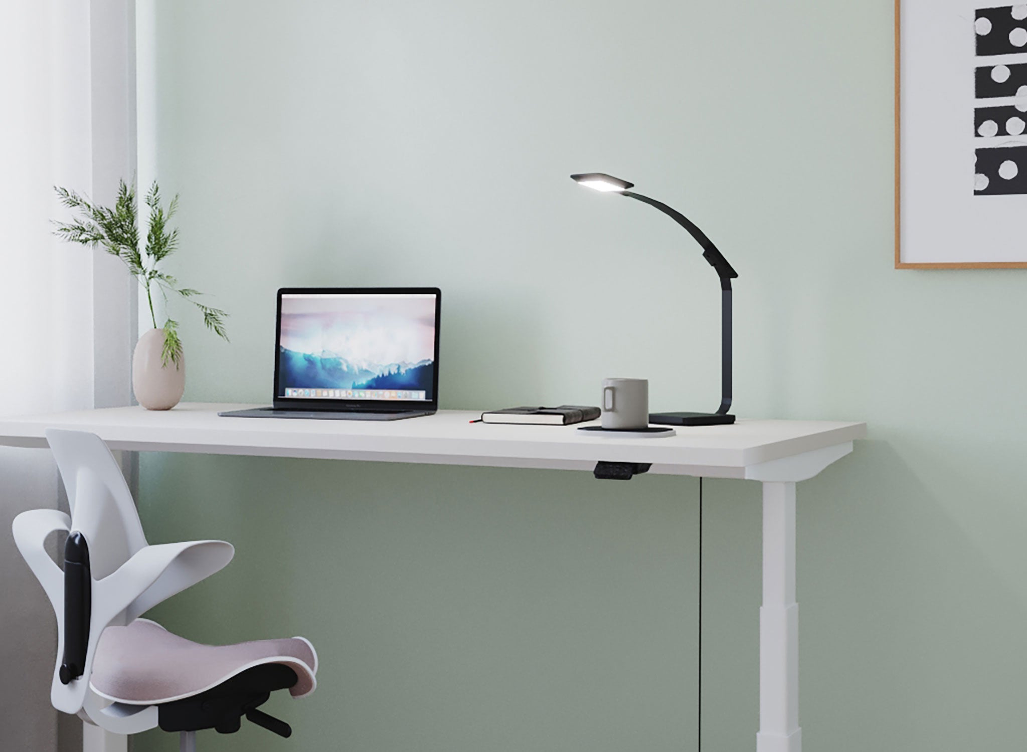 Vor einer hellgrünen Wand sieht man einen weißen Schreibtisch, der elektrisch höhenverstellbar ist. Darauf befinden sich eine schwarze Tischlampe, ein Laptop und eine Pflanze. Daneben steht ein ergonomischer Drehstuhl in Weiß mit taupefarbenem Sitzpolster.