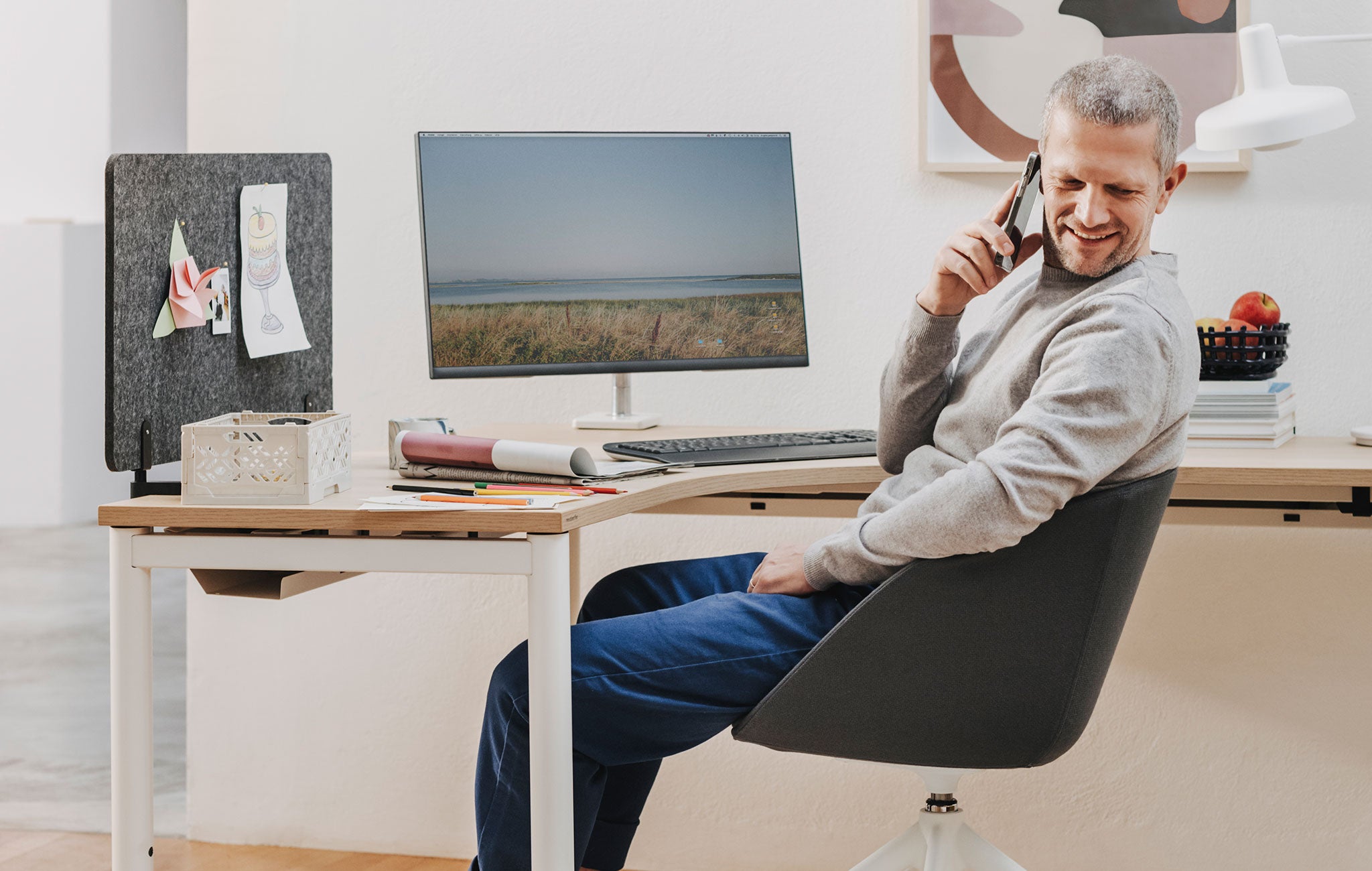 Man sieht einen Raum mit einem Arbeitsbereich, in dem ein Eckarbeitstisch mit Holztischplatte steht. Auf diesem befindet sich ein Monitorscreen mit einer Halterung. Davor sitzt ein älterer Mann, der in der rechten Hand ein Handy zum Ohr hält und nach hinten blickt.