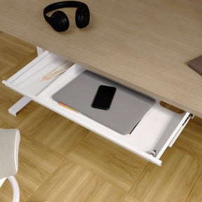 Detailansicht einer weißen Tischschublade. Die Lade ist an einer Tischplatte aus Eiche befestigt. In der Lade ist links ein kleines Fach mit Bleistiften zu sehen. Im rechten, größeren Fach befinden sich ein Laptop und ein Smartphone.
