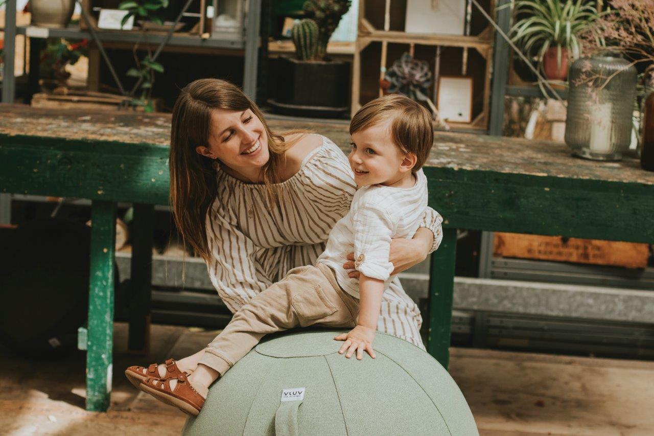 Ein Kind sitzt auf einem grünen Sitzball, während es von einer Frau gestützt wird.
