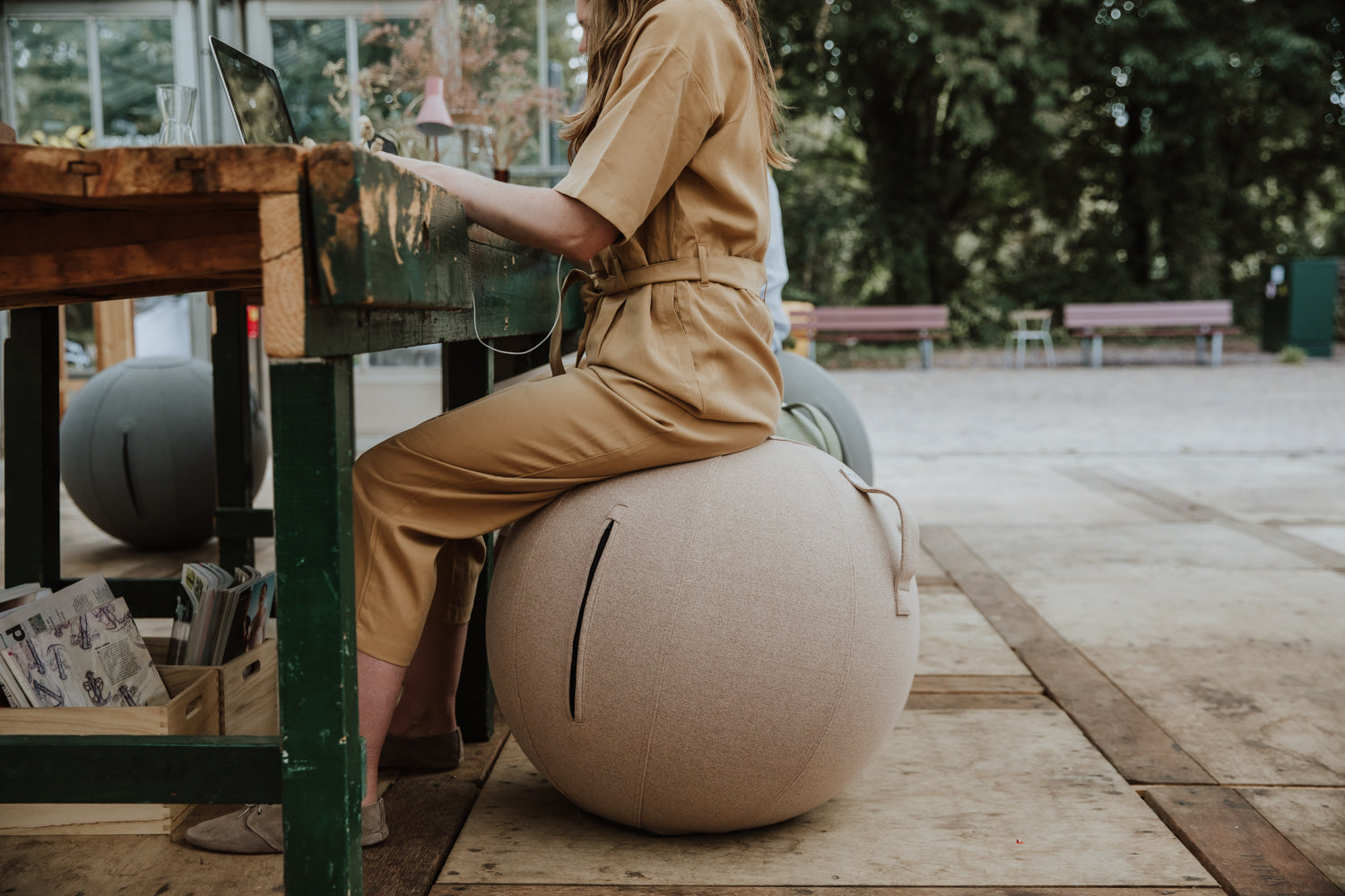 Eine Frau sitzt auf einem beigefarbenen Sitzball und arbeitet mit ihrem Laptop an einem Gartentisch.