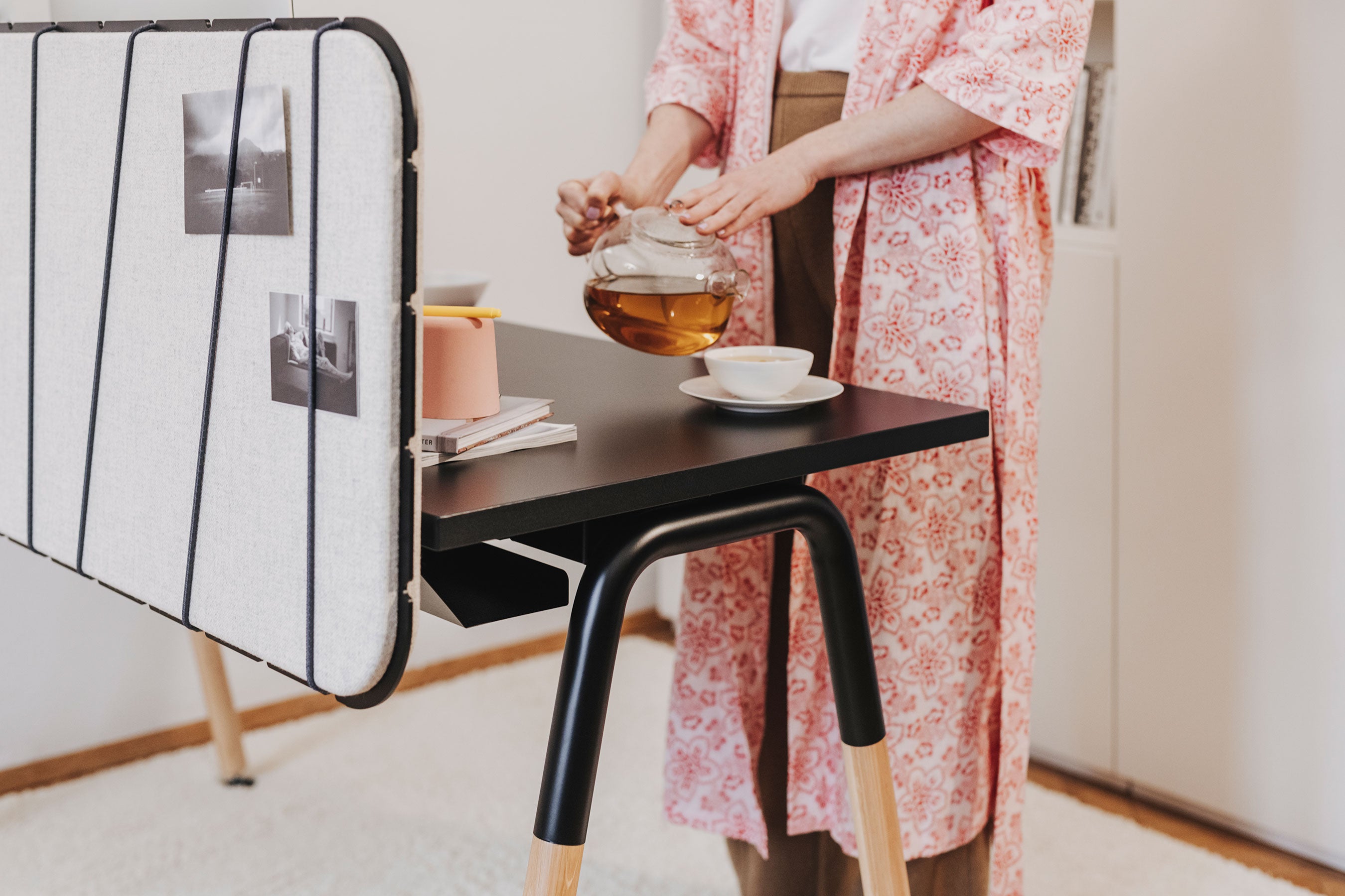 Man sieht in Nahaufnahme einen Arbeitsplatz mit einem schwarzen Arbeitstisch, der Tischbeine aus Holz hat und die Pinnwand ist in grau mit Flexbändern an den Tisch fixiert. Auf ihr befinden sich zwei Fotos. Hinter dem Tisch steht eine Frau und schenkt sich Tee in ein weißes Häferl. Sie trägt einen rosafarbenen floralen Kimono.