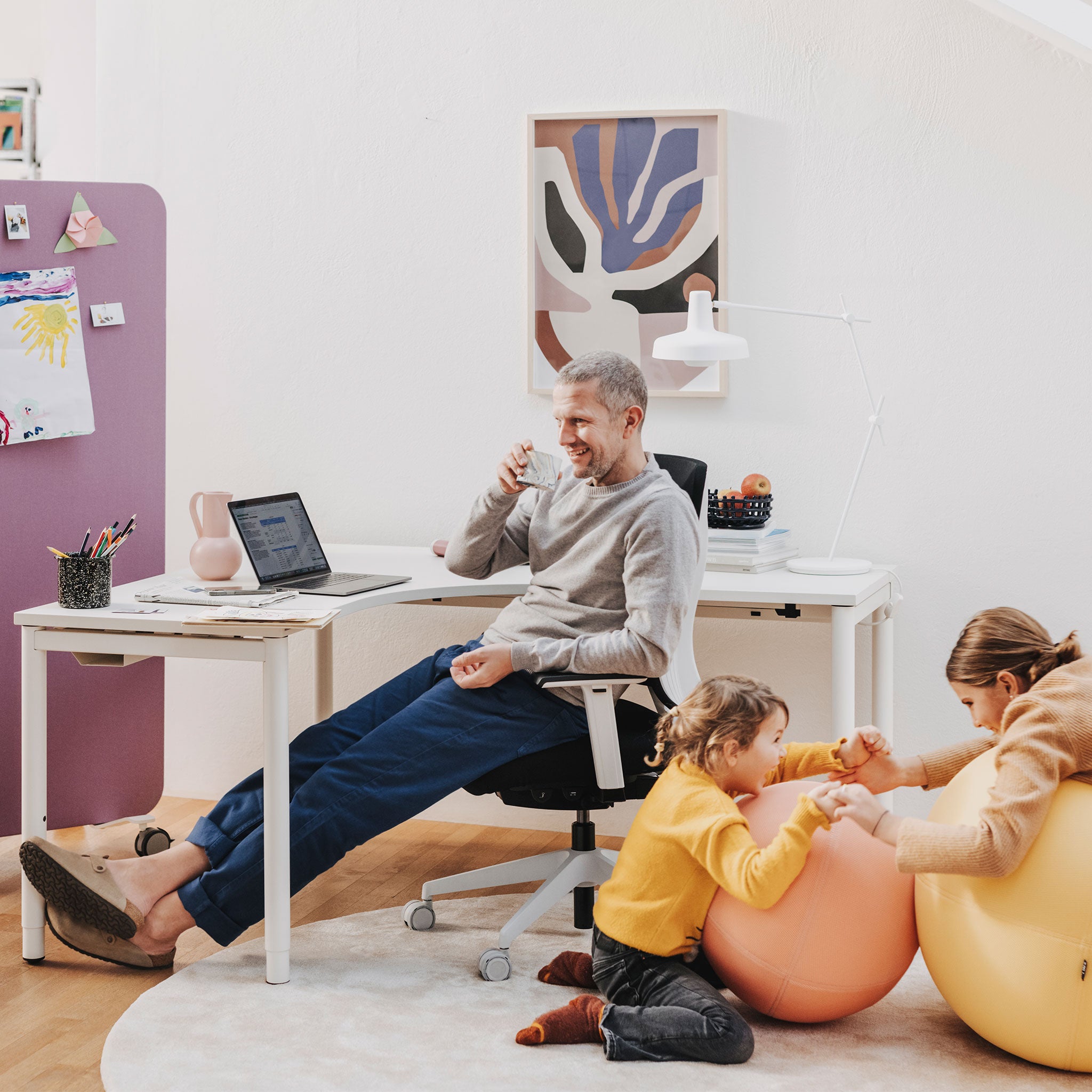 Man sieht einen Raum mit einem Arbeitsbereich, in dem ein Eckarbeitstisch mit Holztischplatte steht. Auf diesem befindet sich ein Laptop und Arbeitsutensilien. Davor sitzt ein älterer Mann, der in der rechten Hand ein Glas zum Mund hält. Rechts neben ihm spielen zwei Kinder auf zwei Sitzbällen.