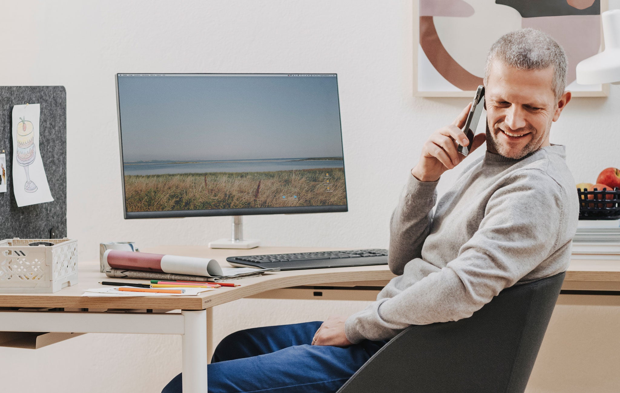 Auf einem Eckarbeitstisch mit einer Arbeitsplatte aus Holz befindet sich ein Monitorscreen mit einer Halterung. Davor sitzt ein älterer Mann, der in der rechten Hand ein Handy zum Ohr hält und nach hinten blickt.