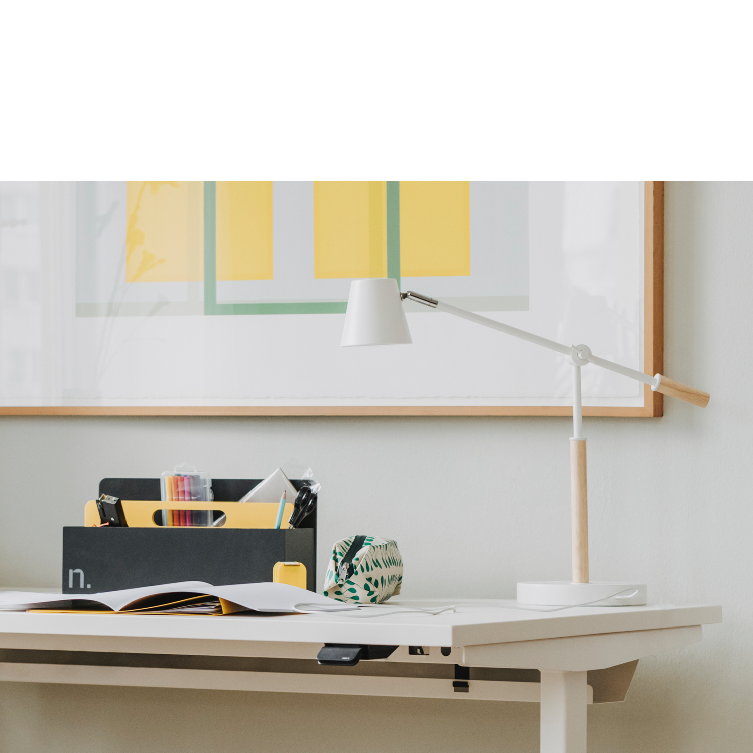 Auf einem weißen Sitz-Steh-Arbeitstisch steht eine Toolbox in graphit mit brillantgelbem Griffelement. In der Toolbox befinden sich Büroutensilien und mehrere Stifte. Auf dem Tisch liegen zwei aufgeschlagene Magazine