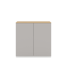 Vor weißem Hintergrund sieht man ein kiesgraues Sideboard mit zwei Drehtüren. Die Türen haben keine Griffe und sind mit einem Push-to-open-Mechanismus ausgestattet. Auf dem Schrank ist eine Abdeckplatte aus Eichenholz angebracht.