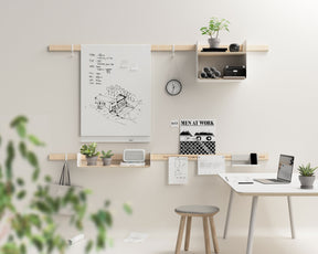 An einer weißen Wand hängt ein Wandregalsystem bestehend aus zwei Holzleisten. An den naturfarbenen Holzleisten sind Haken, ein Whiteboard und Regalfächer angebracht. Davor steht ein Schreibtisch, auf dem ein Laptop und eine Topfpflanze abgestellt sind, sowie ein Hocker mit grauem Bezug.