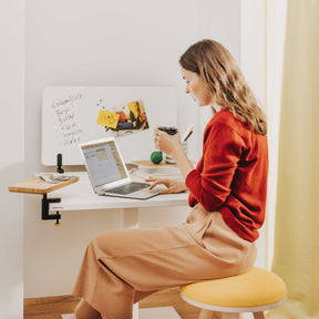 Eine junge Frau mit braunen, langen Haaren und einem roten Pullover sitzt auf einem gelben kleinen Hocker. Vor ihr steht ein weißer Move Mini Arbeitstisch. Auf dem Tisch befinden sich ein Laptop und ein Stapel Unterlagen. Auf dem Tisch ist mit einer schwarzen Klemme ein rechteckiges Bambus-Tablett und ein kleines Whiteboard befestigt.