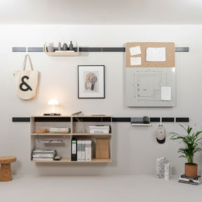 An einer weißen Wand hängt ein Wandregalsystem bestehend aus schwarzen Holzleisten. An den Leisten sind Haken, eine Pinnwand mit Whiteboard und mehrere Regale angebracht. 
