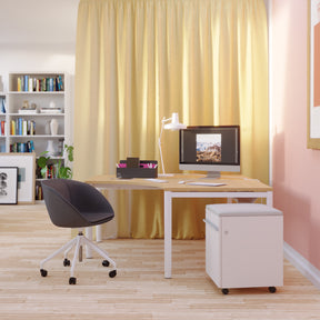 Man sieht einen Raum mit Holzfußboden, in dem sich ein Arbeitsbereich befindet, in dem ein Eckarbeitstisch mit cremefarbener Tischplatte steht. Dahinter ist ein hellgelber Vorhang und auf der rechten Seite ist die Wand rosafarben. Vor dem Tisch steht ein Drehstuhl in schwarz und rechts ein weißer Rollcontainer mit hellgrauem Sitzpolster. Auf dem Tisch steht eine Toolbox und ein Stand-PC und eine weiße Tischleuchte. Im Hintergrund sieht man Bücherregale.