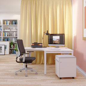 Man sieht einen Raum mit Holzfußboden, in dem sich ein Arbeitsbereich befindet, in dem ein Eckarbeitstisch mit cremefarbener Tischplatte steht. Dahinter ist ein hellgelber Vorhang und auf der rechten Seite ist die Wand rosafarben. Vor dem Tisch steht ein Drehstuhl in schwarz und rechts ein weißer Rollcontainer mit hellgrauem Sitzpolster. Auf dem Tisch steht eine Toolbox und ein Stand-PC und eine weiße Tischleuchte. Im Hintergrund sieht man Bücherregale.