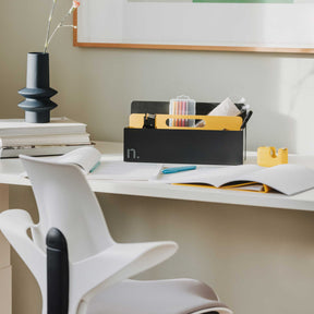Auf einem weißen Sitz-Steh-Arbeitstisch Move steht eine Toolbox in graphit mit brillantgelbem Griffelement. In der Toolbox befinden sich Büroutensilien und mehrere Stifte. Auf dem Tisch liegen zwei aufgeschlagene Magazine sowie ein Bücherstapel, auf dem sich in einer dunkelgrauen Vase Nelken befinden.