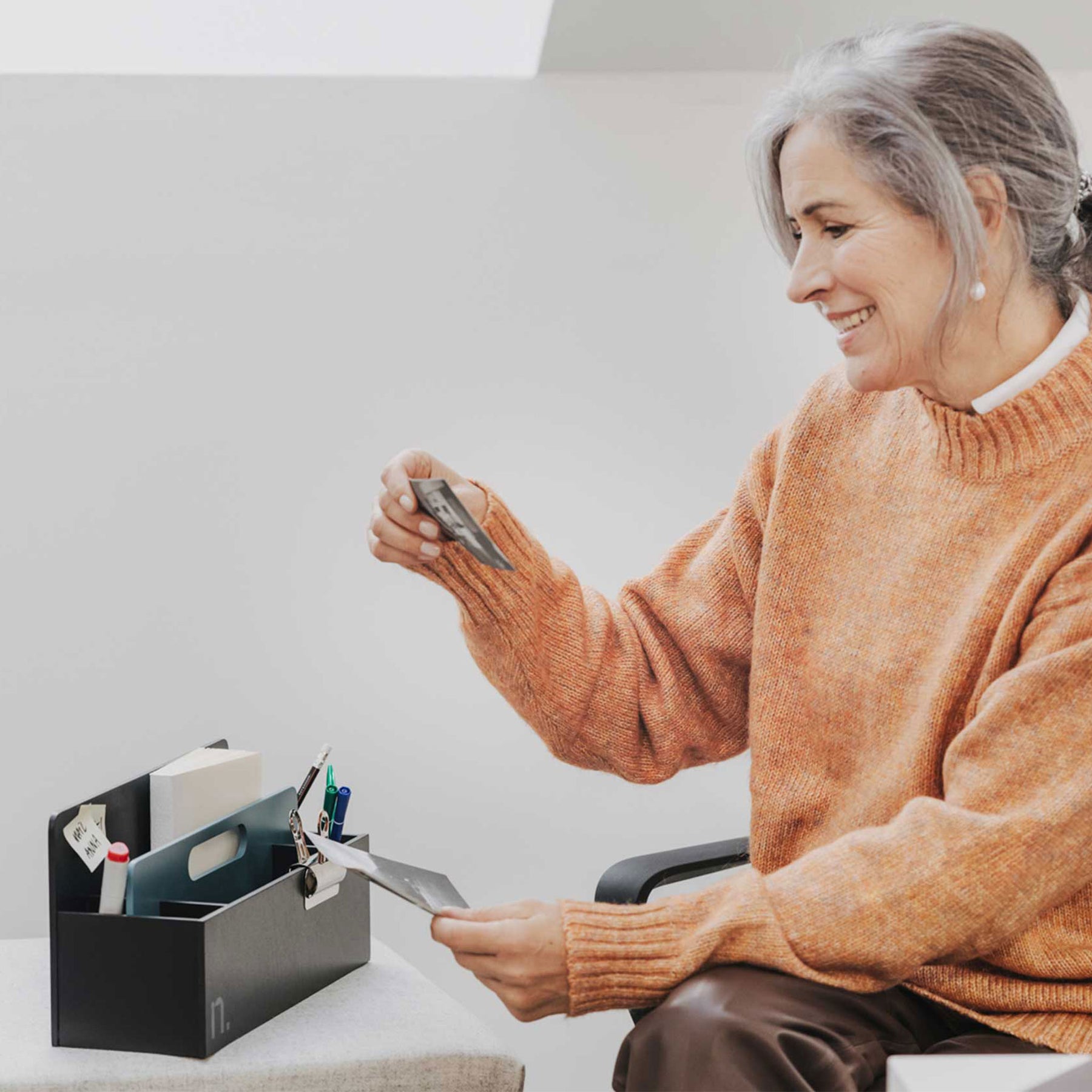 Eine ältere Frau sitzt neben einem Rollcontainer mit hellgrauem Sitzpolster. Die Frau hat graumelierte Haare und trägt einen orangen Pullover. Sie hält in beiden Händen jeweils ein Foto. Auf dem Rollcontainer steht eine Toolbox in graphit mit niagaragrünem Griffelement.