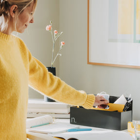 Eine Frau mit blonden Haaren und gelbem Pullover greift nach einer Toolbox, die auf einem Arbeitstisch steht. Die Toolbox ist graphit mti einem Griffelement in brillantgelb. In der Toolbox sind eine Schere, Stifte und ein Notizbuch verstaut. Auf dem Tisch befinden sich ein aufgeschlagenes Buch, mehrere Stifte, ein Bücherstapel und eine graue Vase mit lachsfarbenen Nelken.