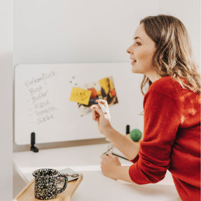 Eine Frau mit braunen Haaren und rotem Pulli schreibt eine Einkaufsliste auf ein Whiteboard, das mittels zwei Multiklemmen am cremeweißen Arbeitstisch montiert ist. LIinks vorne ist ein Bambus-Anbautablet am Tisch montiert, darauf steht eine gesprenkelte Kaffeetasse.